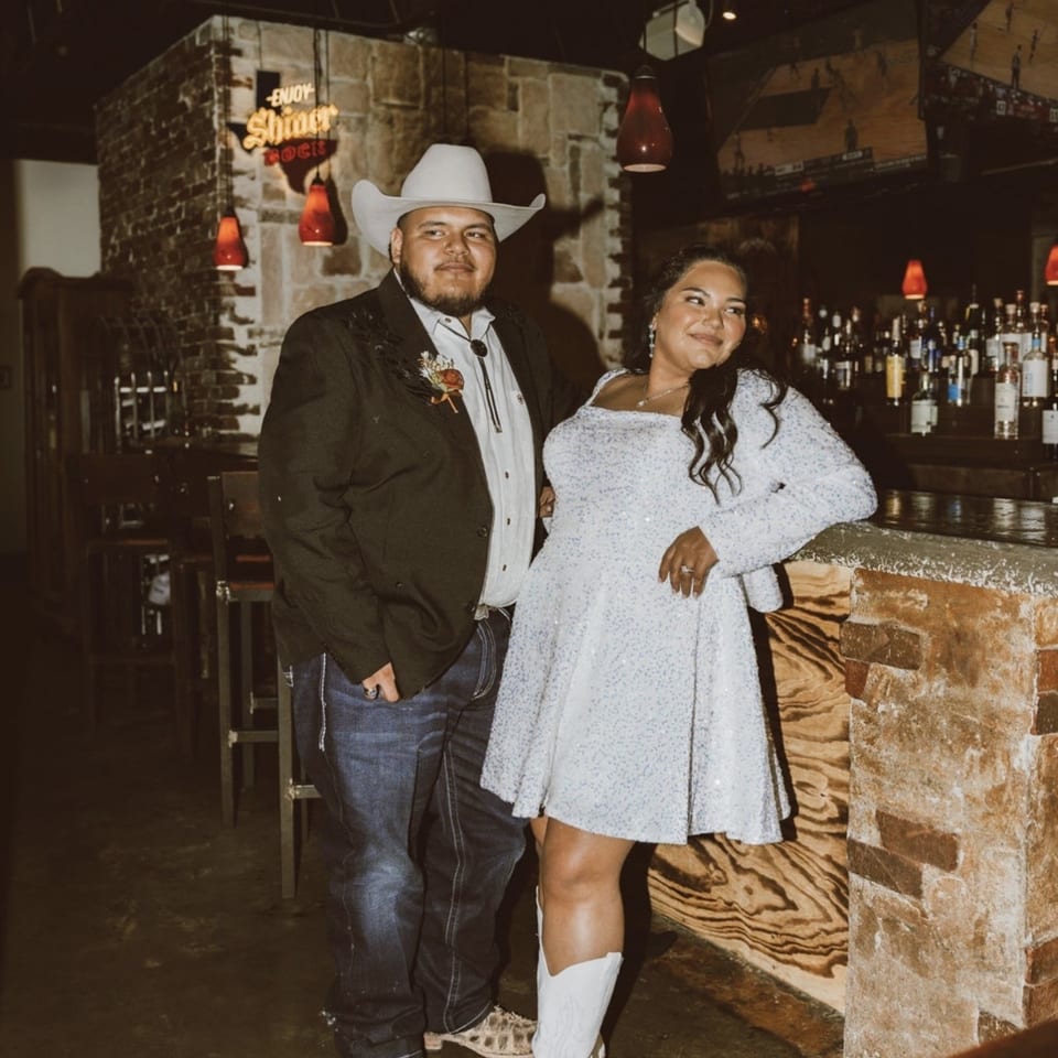 Two people pose inside a rustic bar, wearing western-inspired outfits beside a brick counter and illuminated liquor shelves.