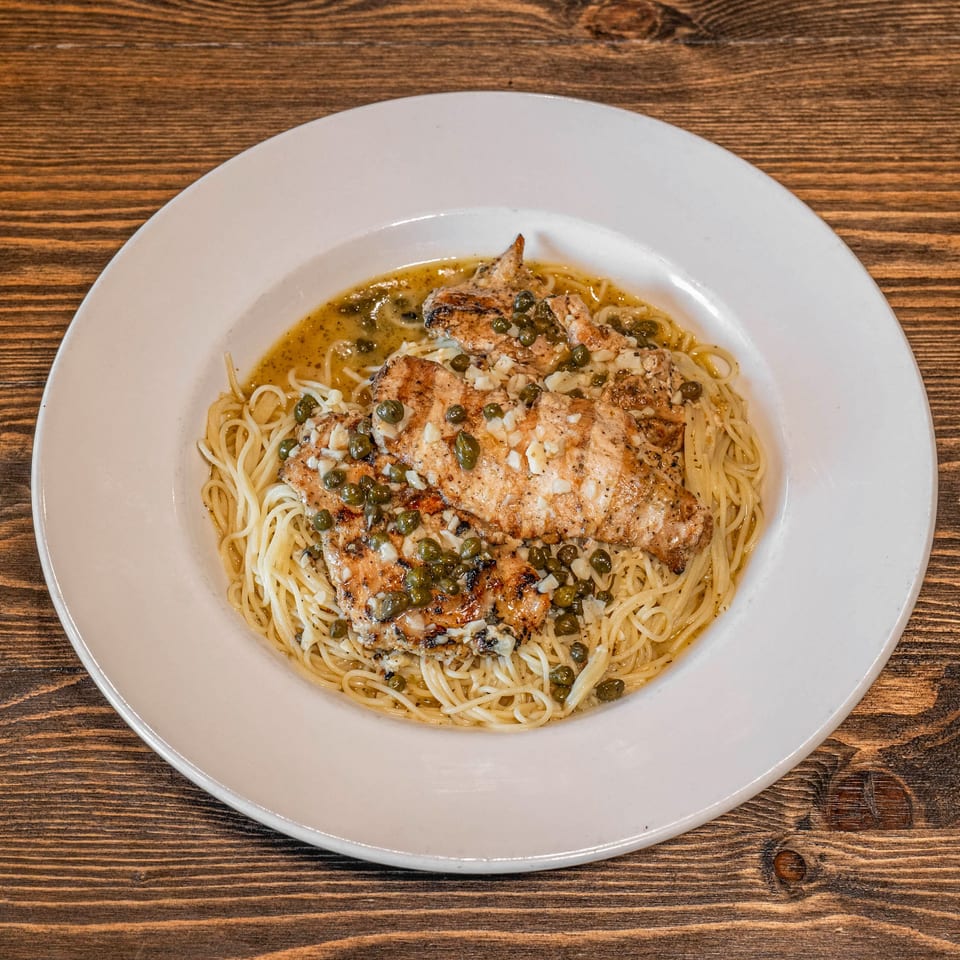 Grilled chicken piccata with capers and diced garlic served over spaghetti in a white plate on a wooden table.