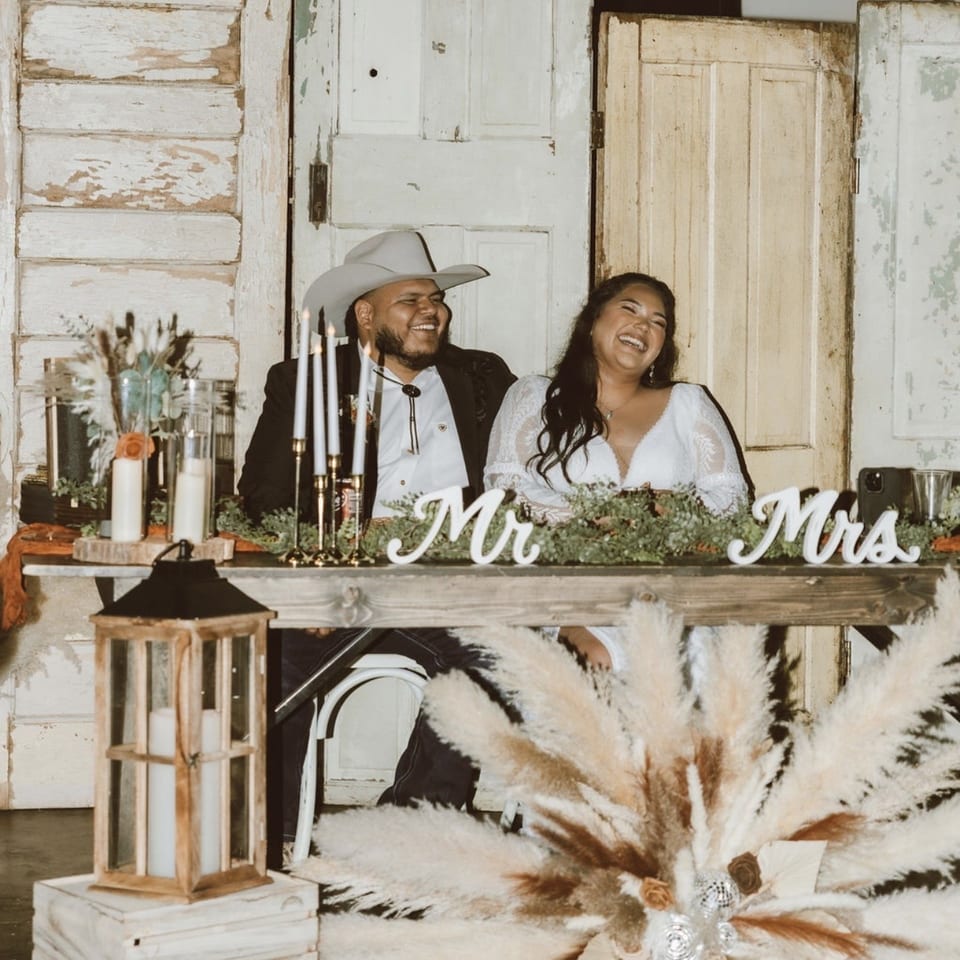 Newlywed couple laughing behind a rustic wedding table decorated with candles, greenery, pampas grass, and “Mr” and “Mrs” signs.