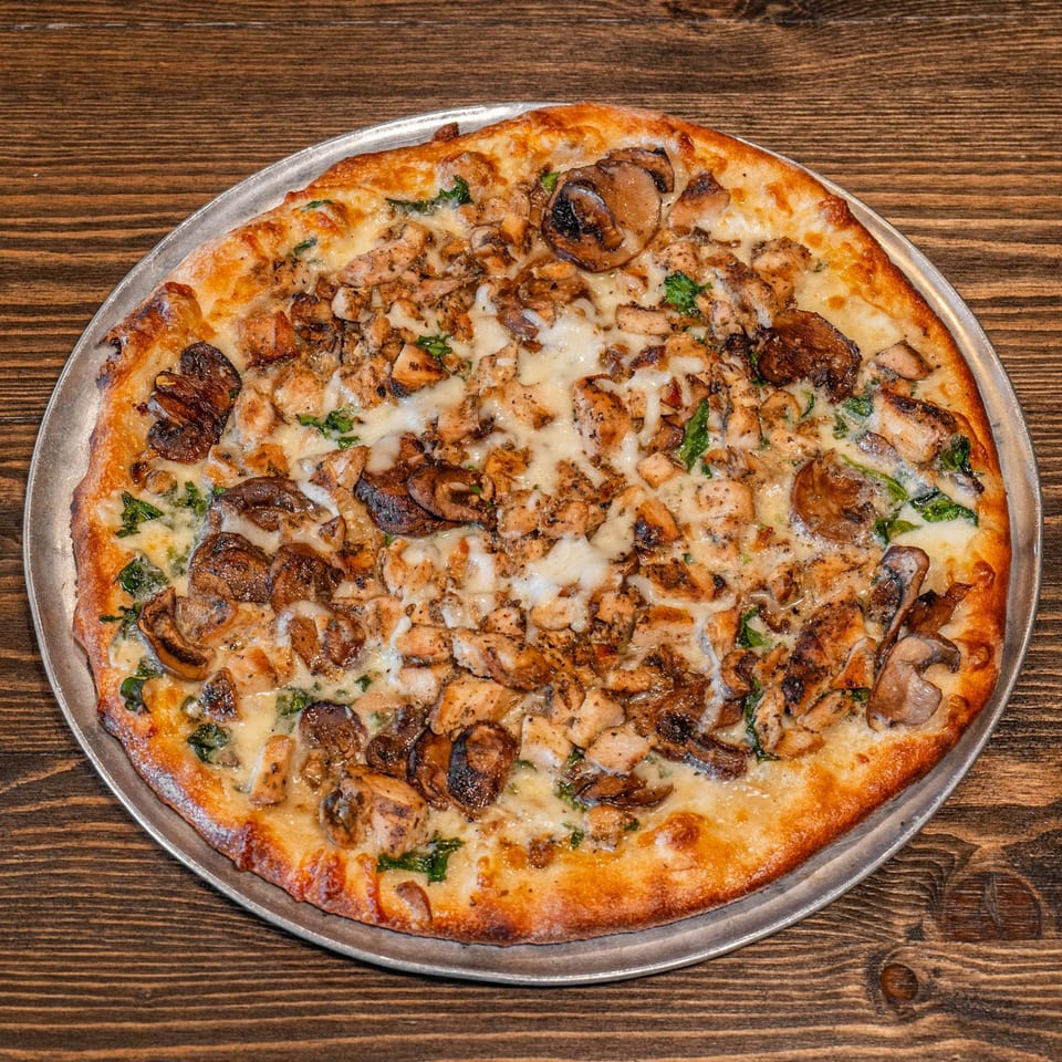 Cheesy pizza topped with grilled chicken, mushrooms, and greens on a metal pan atop a rustic wooden table.