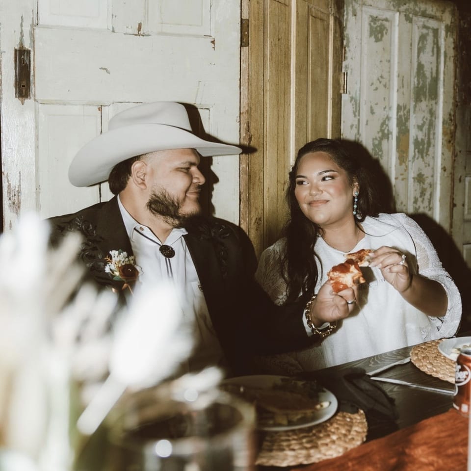 Couple dining at a rustic restaurant table, smiling at each other while the woman holds a slice of pizza.