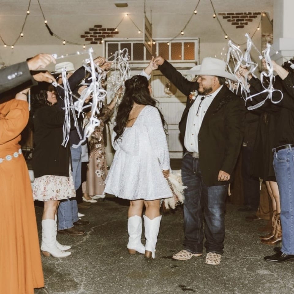 Wedding guests form an arch with white streamers as a couple dances beneath hanging string lights indoors.