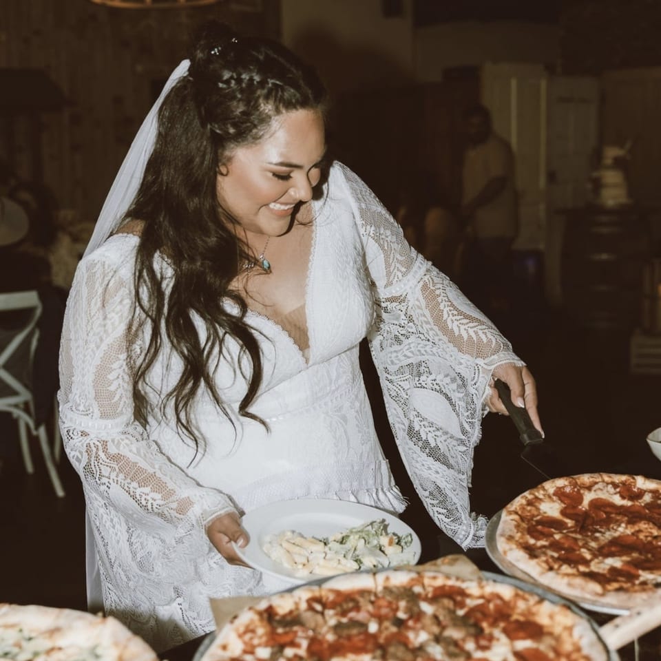 Smiling bride in a lace dress serves pasta beside several pizzas in a warmly lit restaurant.