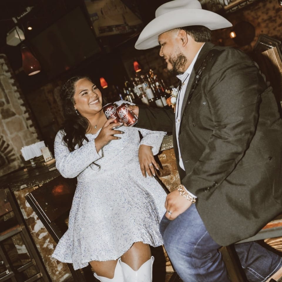Smiling couple in Western attire pose inside a dimly lit restaurant, holding Dr Pepper cans near the bar.